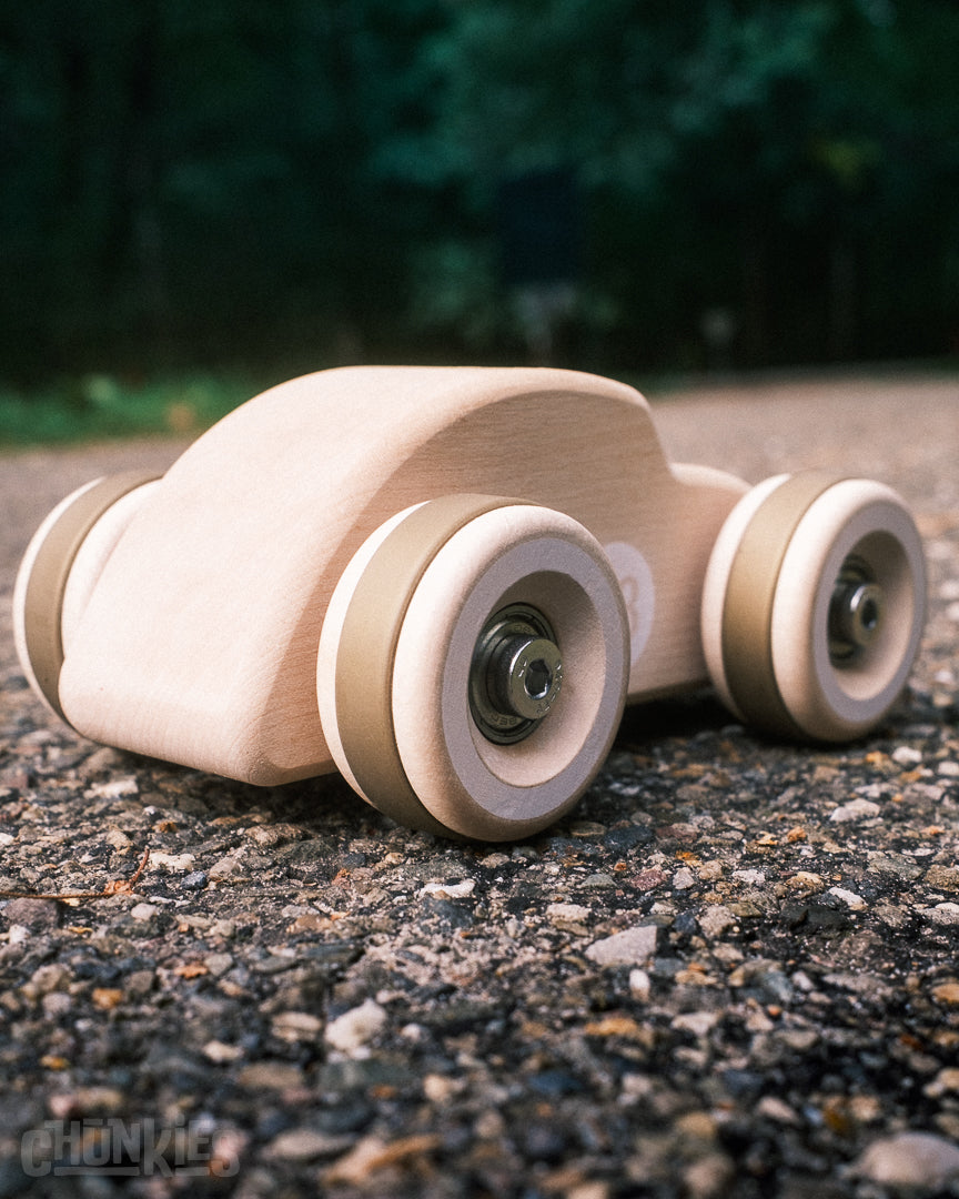 Chunkies' natural solid maple wooden toy car in the shape of a classic beetle with big wooden wheels, real skate bearings and stainless steel hardware on a road surface and a forest in the background.